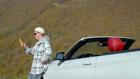 hombre usando el teléfono al lado de un coche descapotable blanco en las montañas