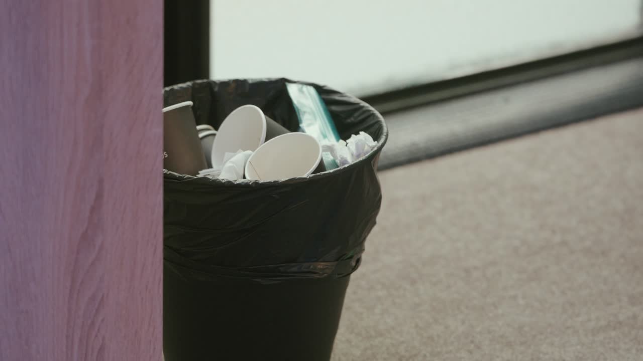 Papelera negra con vasos desechables y pañuelos en un suelo enmoquetado.