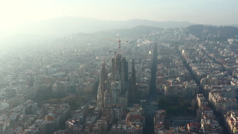 Aerial-footage-of-basilica-Sagrada-Familia.-Large-unfinished-church-with-tower-cranes.-Hazy-view-against-sun.-Barcelona,-Spain