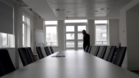 an individual pacing up and down in office conference room, passing by a lengthy table encircled by chairs, talking on mobile one