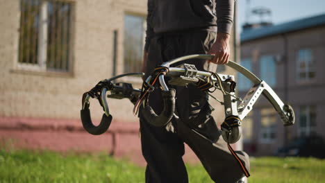 person holding metallic stilts with dark grips and straps in grassy outdoor , wearing dark gray hoodie and loose pants, only hands and lower body visible, background blurred with grass and building