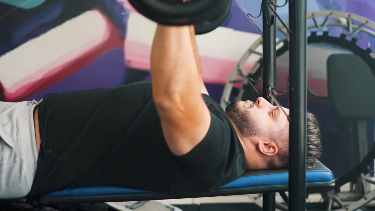 Genç bir adamın spor salonunda halterle bench press antrenmanı yaparken yan görünüm, hareketli çekim.