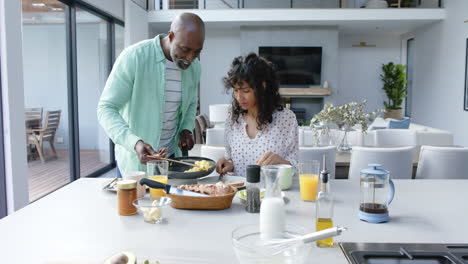una pareja bi-racial feliz desayunando con huevos revueltos en la cocina, en cámara lenta.