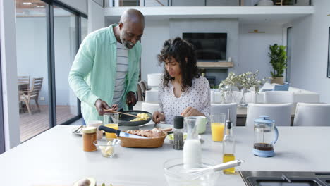 happy biracial couple having breakfast with scrambled eggs in kitchen, slow motion