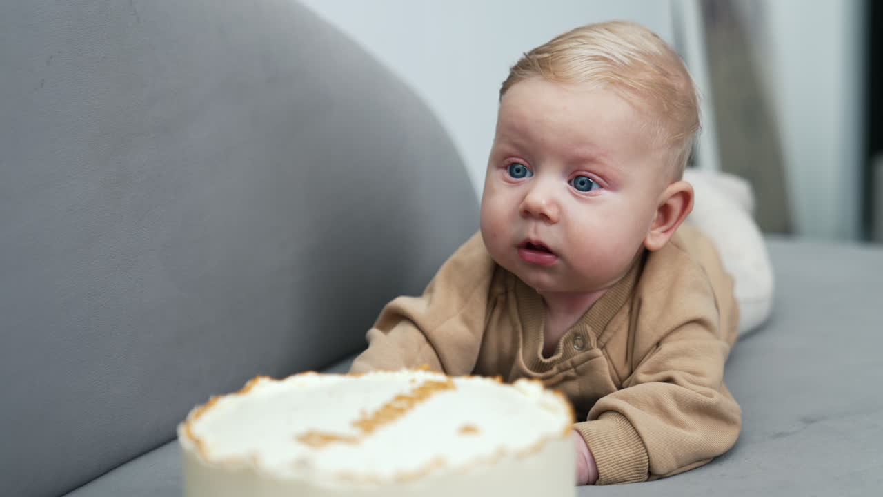 Beautiful blue-eyed blond infant child lies on sofa. A big cake is in front of a cute baby boy.