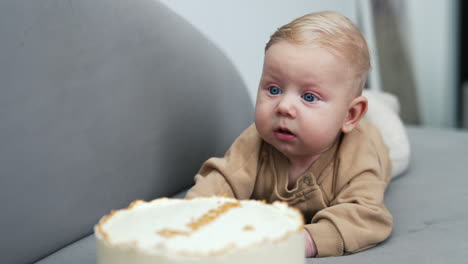 beautiful blue-eyed blond infant child lies on sofa. a big cake is in front of a cute baby boy.