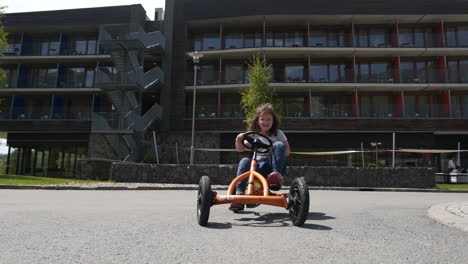 rotating fhd shot of a small girl cycling on an orange pedal quadricycle in a private resort in dolní morava, czechia with her family cycling behind