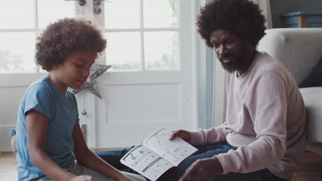 middle aged black father sitting on the floor at home helping his pre teen son with the instructions for a toy construction kit, close up, low angle, side view