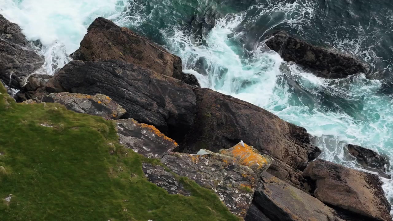 Aerial footage showing rocky cliffs with green grass and ocean waves crashing along the coastline in a natural outdoor setting.