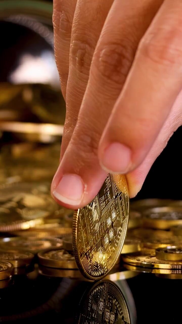 Una mano in primo piano posiziona una moneta di criptovaluta dorata in un barattolo di vetro, superficie nera riflettente, illuminazione drammatica.