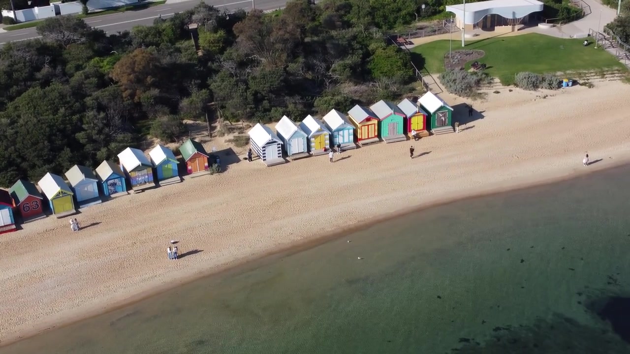 Vista aerea con drone di colorate capanne sulla spiaggia che costeggiano la riva di ciottoli, con persone che camminano vicino all'acqua calma e alberi lungo il lungomare a Brighton, Inghilterra, Regno Unito