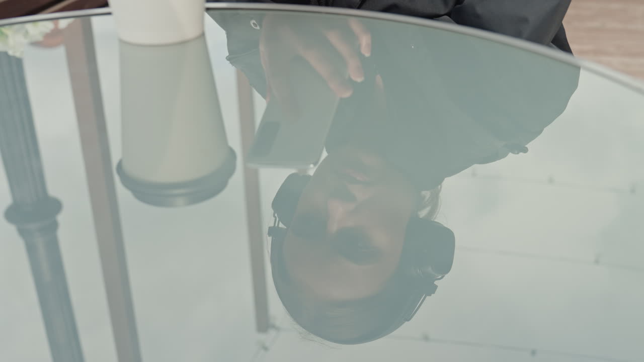 Mulher pensativa de fones de ouvido em um café ao ar livre, bebendo café ao lado de uma mesa de vidro que reflete o céu nublado, navegando em um smartphone, usando um casaco escuro e fones de ouvido grandes, terraço silencioso em uma cidade chuvosa, reflexiva