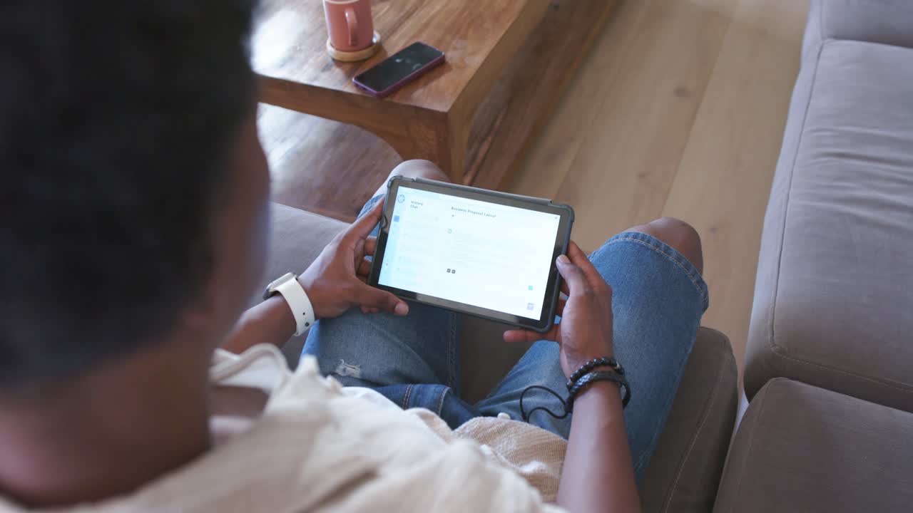 African American man settling on sofa with tablet, reading and scrolling with wired earbud plugged