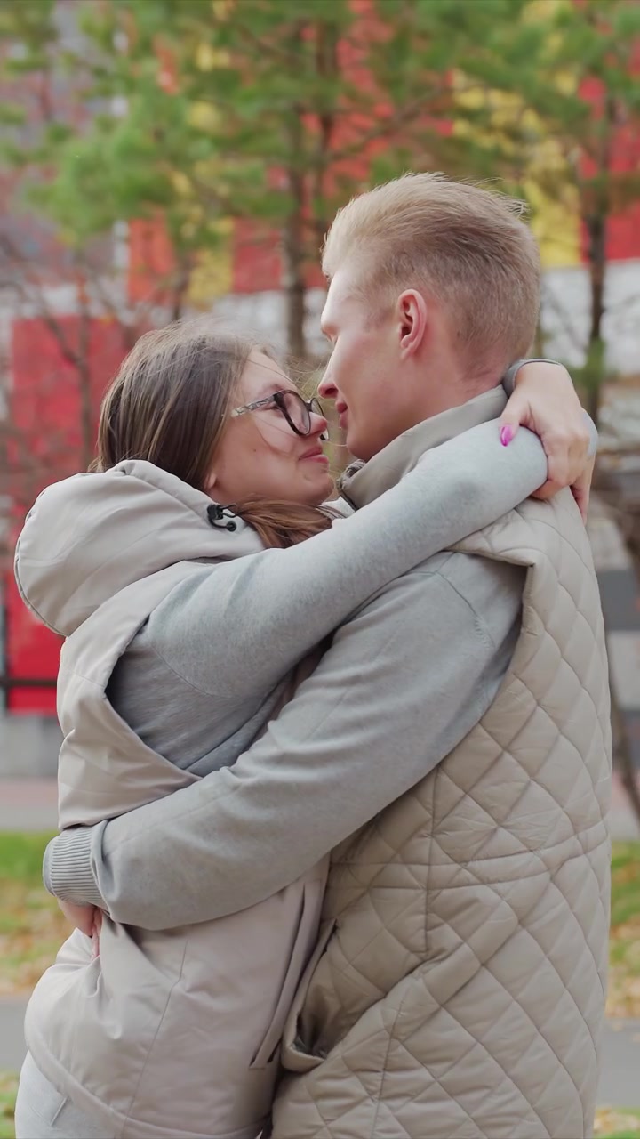Una pareja se abraza estrechamente al aire libre durante el otoño. La pareja mantiene contacto visual o se mira mutuamente. El suelo muestra follaje de otoño caído, sugiriendo un cambio estacional.