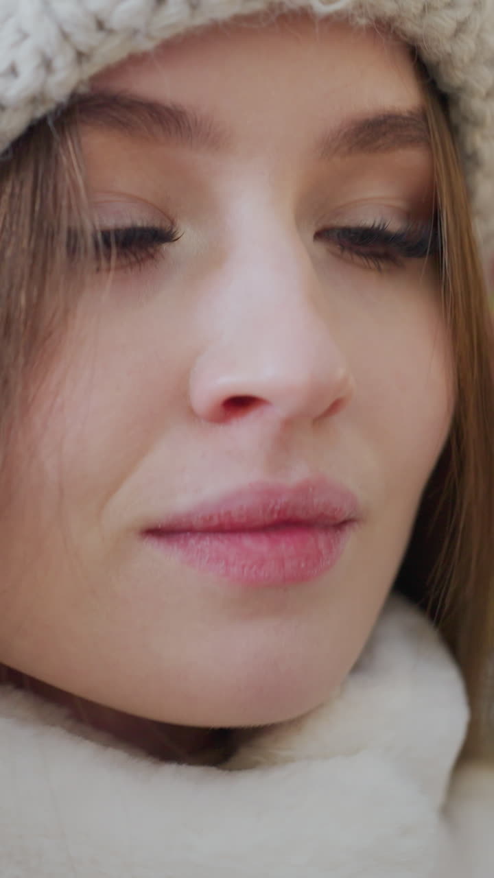 Close-up of woman in fluffy winter jacket adjusting white beanie with blurred background featuring people and festive decor, cozy winter fashion moment