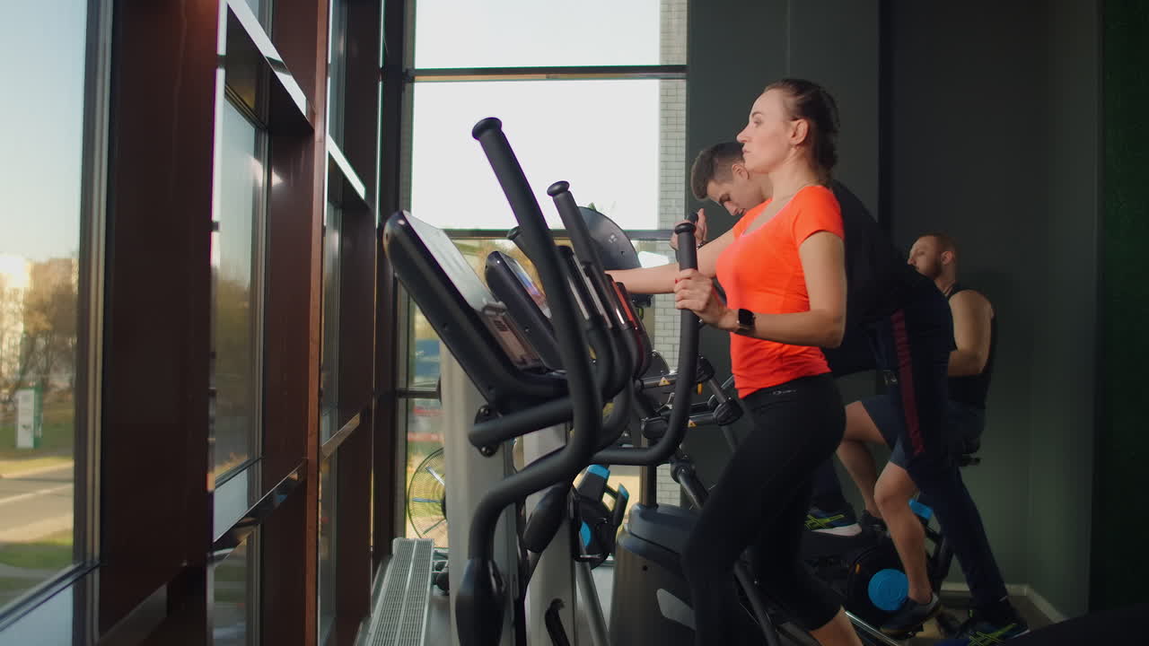 A group of people in the cardio area of the fitness room. Man uses woman's ellipsoidal exercise bike. Young fit woman using an elliptic trainer in a fitness center. A group of young women train on sports training equipment in a fitness gym.