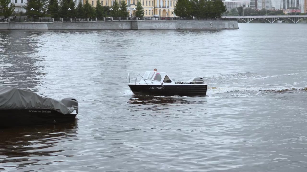 Malý černý motorový člun s jedním dospělým klouže po široké městské řece podél kamenného nábřeží a říční promenády, zvedá bílé kapky, silnou brázdu a vlnky pod zataženou oblohou.