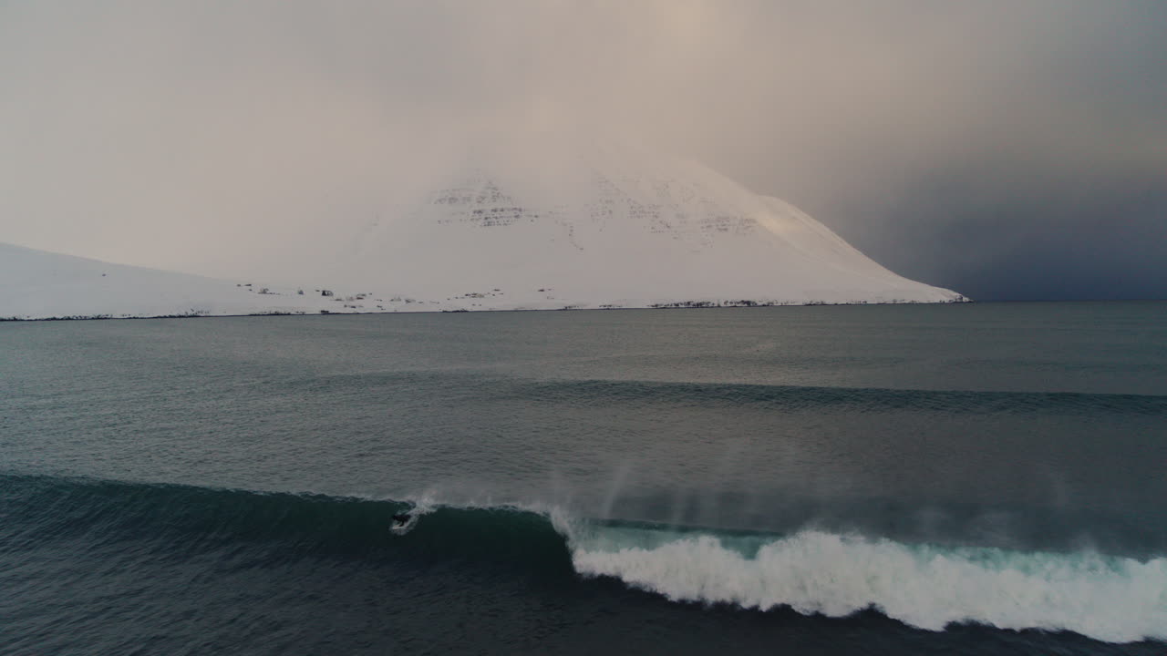 Surfer takse off on wave along snow covered Arctic coastline under dramatic overcast skies with icy water and serene atmosphere