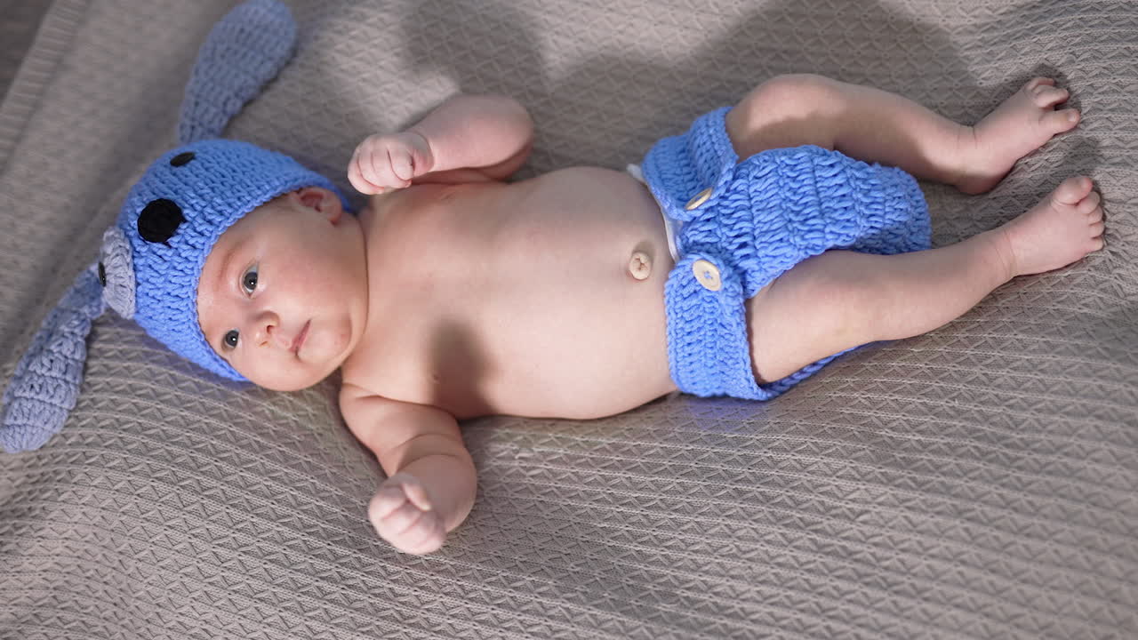 Tiny boy in a blue knitted hat and diaper lies on his back. Funny puppy costume for a small baby. View from above.