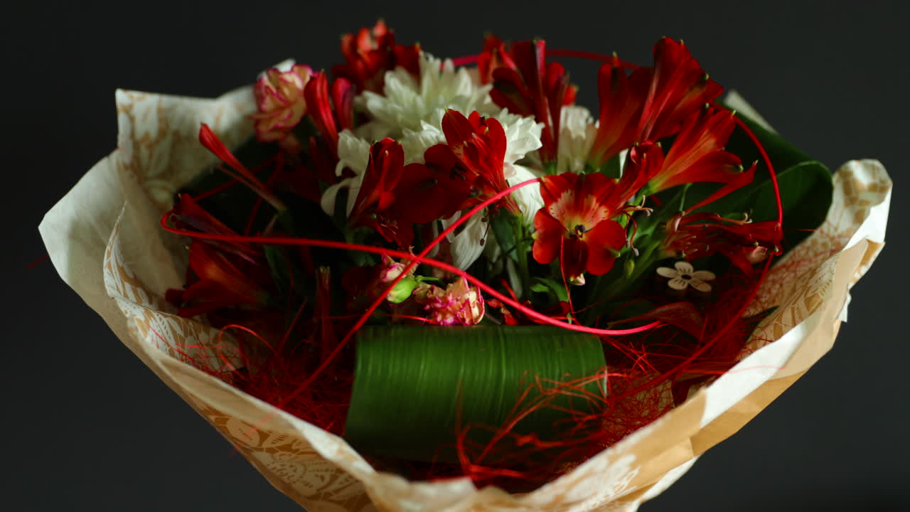 Zoomed in detailed a large bouquet of red and white flowers and green decorative elements in a glass vase decorated with paper around on a dark background