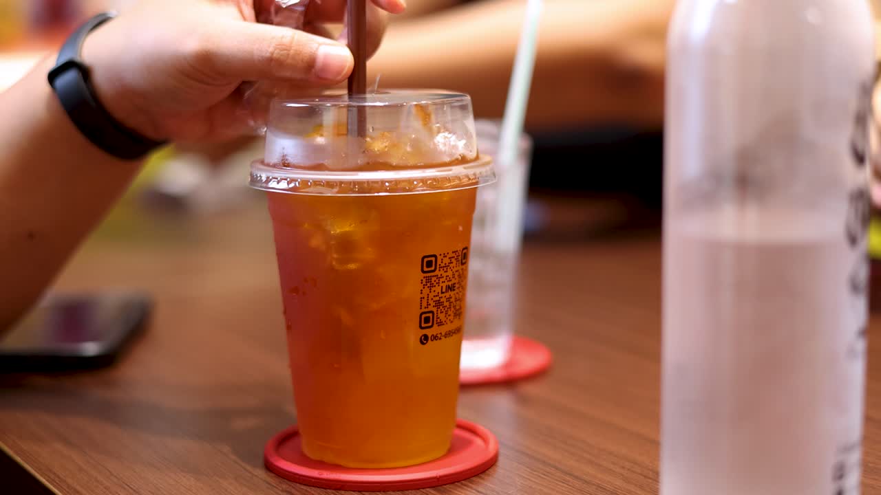 A person stirs iced lemon tea in a clear plastic cup at a wooden café table, with warm indoor lighting and shallow depth of field