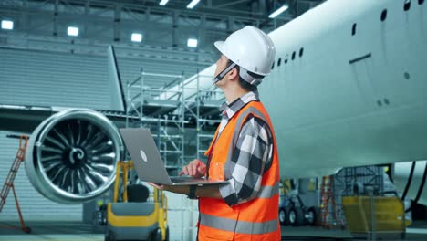 vista lateral de un ingeniero masculino asiático con casco de seguridad mirando una computadora portátil y mirando a su alrededor mientras está de pie con la aeronave en el hangar