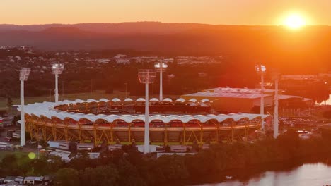 drone glides over a large sports stadium at sunset, capturing dramatic lighting, illuminated field lights, and the adjacent river in gold coast, australia