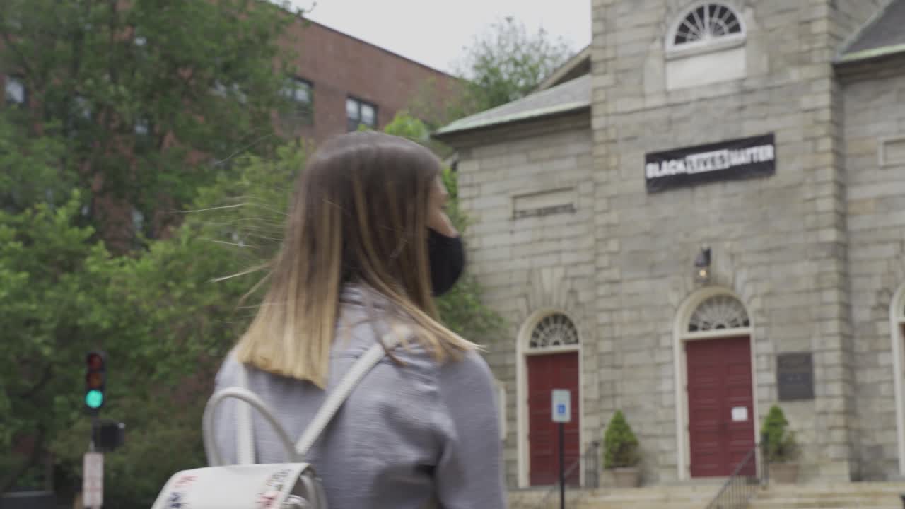 금발 여성은 길을 건너기 전에 왼쪽과 오른쪽을 보고, 검은 얼굴 마스크를 쓰고, 교회 정면에 있는 흑인 생명 문제 현수막을 쓰고, 프레임 밖으로 걸어나가고, 코로나바이러스 대유행, 정적 샷