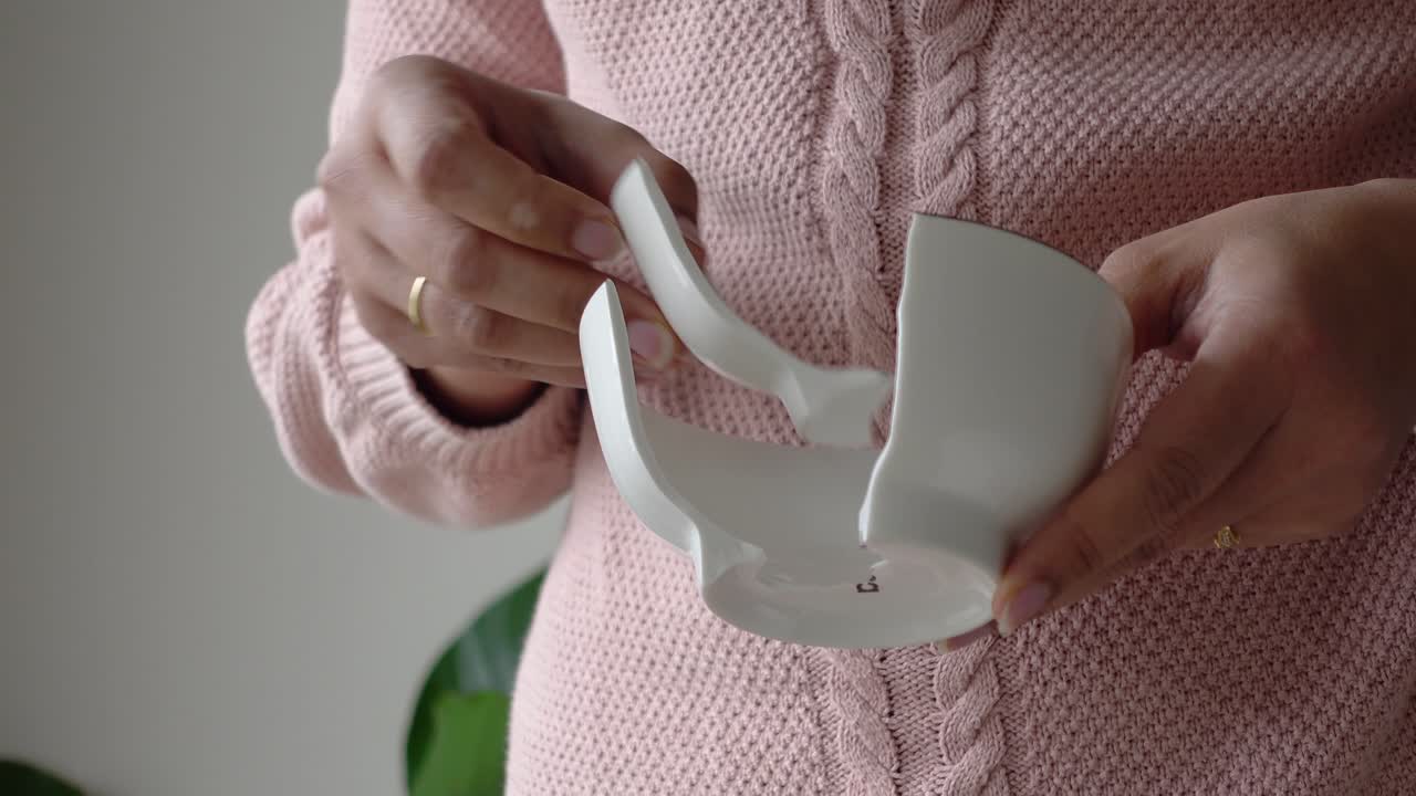 Primo piano di mani che tengono i pezzi di una tazza bianca rotta.