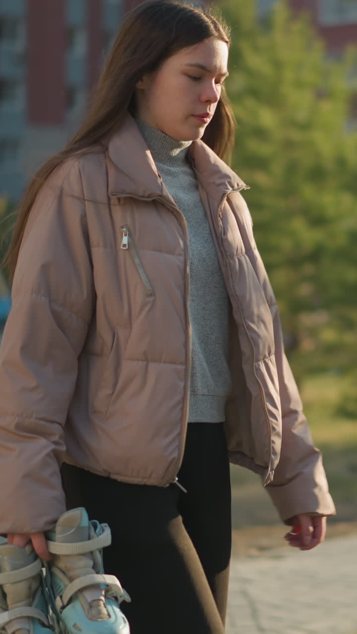 A young girl is walking through a sunlit park, holding a pair of rollerblades in one hand. She is dressed in a peach jacket, grey inner shirt, and black trousers