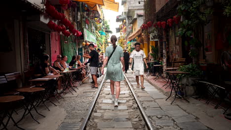 rearview of woman in grey dress walking on train street, narrow path with metal tracks between buildings and tables along edge