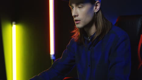 close up view of young serious man wearing cap and playing a game on the computer in a room with colorful neon lamps on the wall