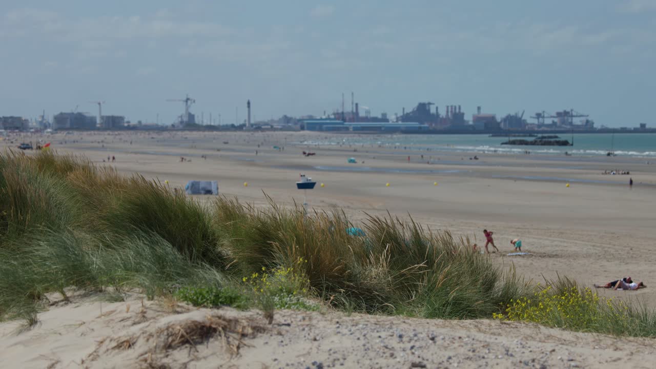Static wide shot of sandy beach, dunes, distant harbor, scattered people, bright daylight, calm mood