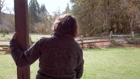 a middle aged woman leaning on a cabin porch thinking and looking off into the forest beyond the field