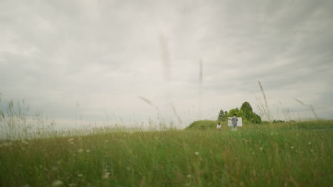 a back view of a painter, wearing a hat and checkered shirt, intently focused on painting on a board in the middle of a tall grass field under a cloudy sky. nearby, a woman sits comfortably on a chair