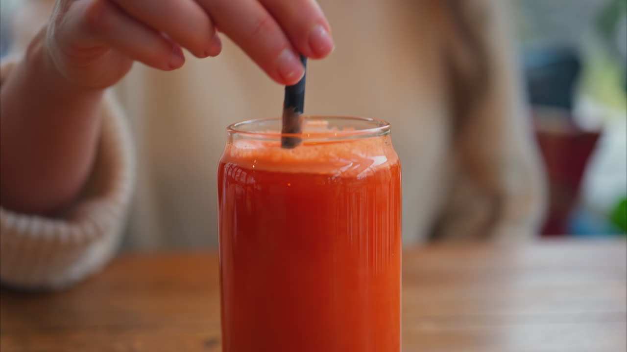 Primo piano di una donna che mescola un succo di arancia e carota in un bicchiere con una cannuccia nera su un tavolo di legno
