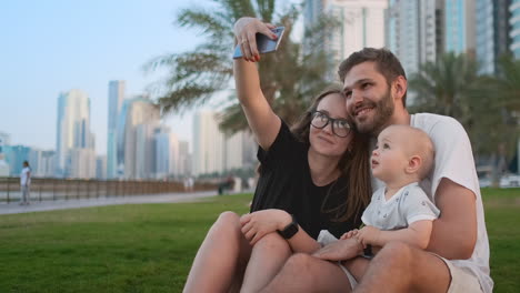 Happy-family-with-two-children-sitting-together-on-grass-in-park-and-taking-a-selfie.-With-smartphone.