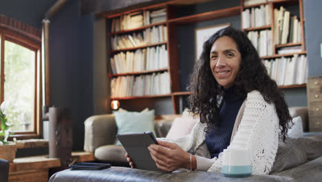 Portrait-of-happy-biracial-woman-sitting-on-sofa-and-using-tablet-at-home,-slow-motion,-copy-space