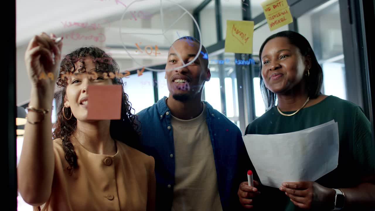 African American team with woman placing pink note, writing on glass with marker, brainstorming