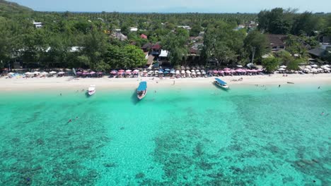 Aerial view of Koh Phi Phi, Thailand beach showing anchored boats, sunbathers, umbrella-lined shore, tropical forest, and resort buildings beside clear turquoise waters