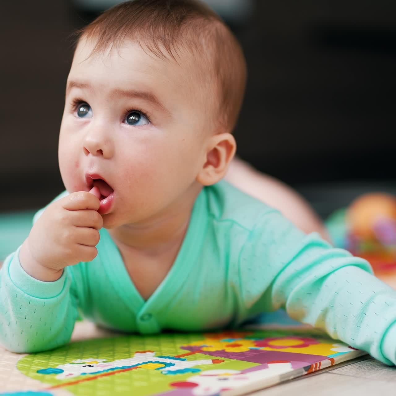Lovely little kid lying on the colorful mat and looking up. Sweet toddler putting his thumb into mouth. Blurred backdrop
