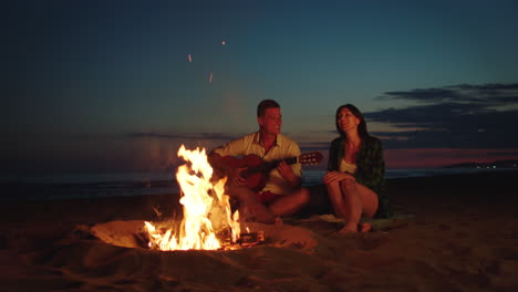 pareja tocando la guitarra junto a una fogata en la playa por la noche