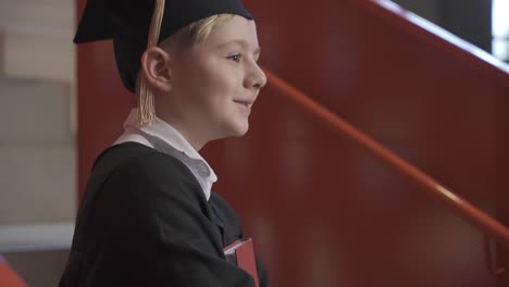 vista lateral de un feliz estudiante de preescolar caucásico con toga y birrete sosteniendo un libro en la ceremonia de graduación