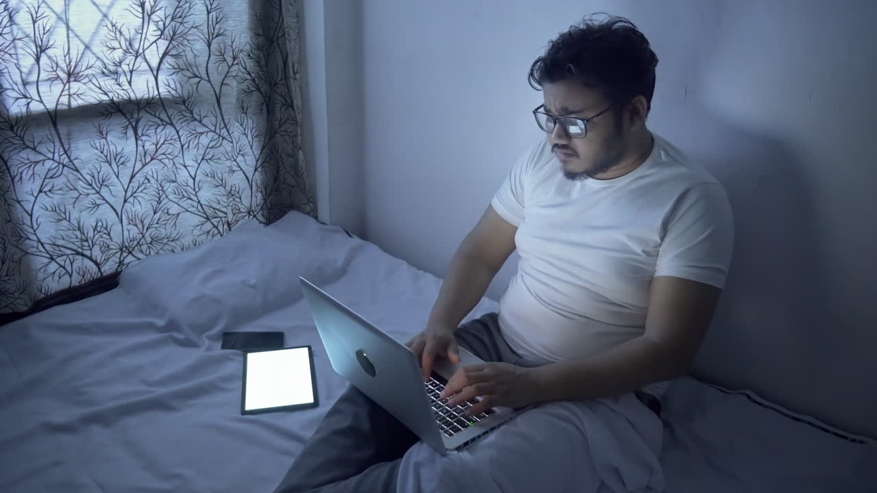 Giovane indiano stressato o stanco dal lavoro, che usa la tecnologia informatica per tutta la notte sul suo letto