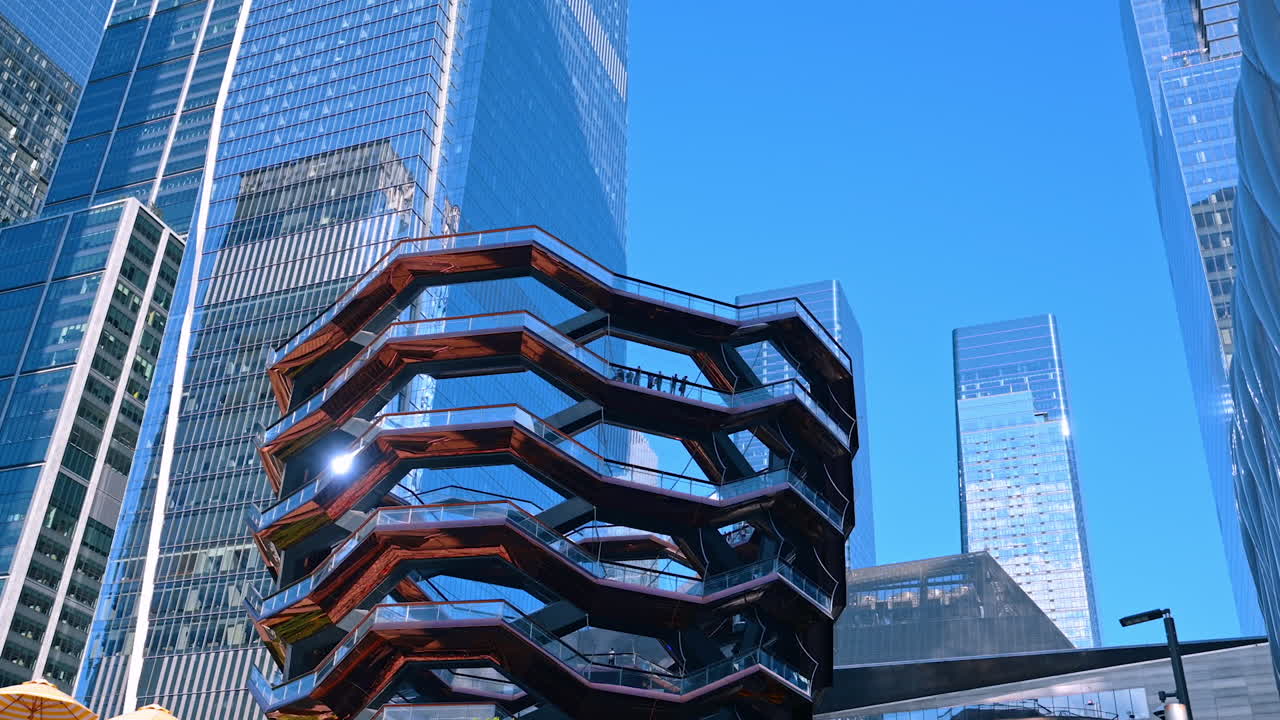 New York, USA, 5 August 2025: People stand on the balcony of the amazing Vessel structure. Stunning glass high-rises are around at the backdrop. Low angle view. New York, USA