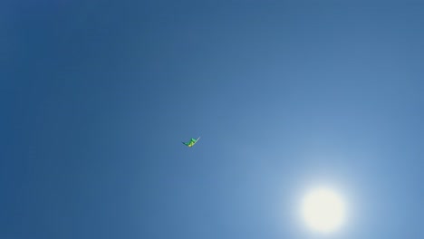 vista de ángulo bajo de la cometa de juguete verde voladora cerca del sol brillante en el cielo azul claro