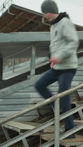 a young boy wearing a gray sweater and a black beanie sprints up and down rusty iron staircase, with the background of metal roofs and surrounding structures