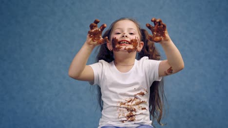cheerful little girl licking chocolate from her fingers. dirty face and white t-shirt of a happy child. funny kid in smeared chocolate isolated on blue.