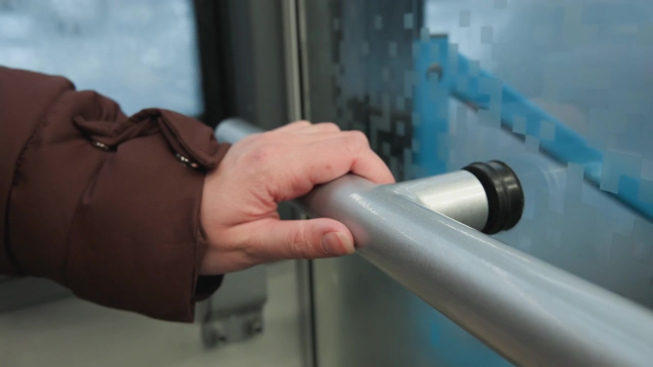 Primer plano de una mano sosteniendo una barandilla tubular de acero inoxidable dentro del transporte público cerca de una ventana salpicada de lluvia, manga de chaqueta marrón visible, agarre firme que transmite equilibrio, comodidad, movimiento.