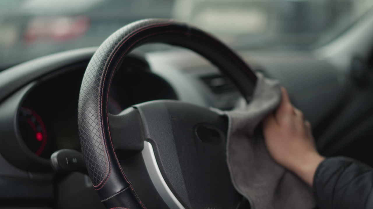 Hands of car enthusiast cleaning steering wheel with microfiber cloth in circular motion inside modern car interior, detail shot showing quilted seat cover and dashboard blur, in bright daylight
